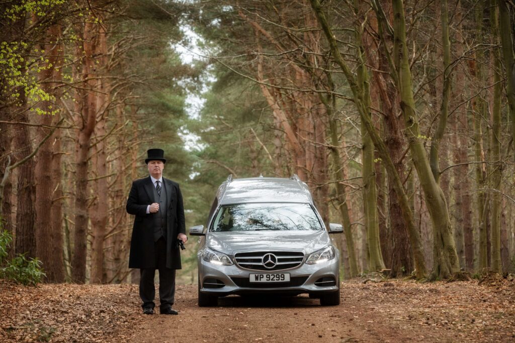 Photo of a funeral director standing beside a modern Mercedes hearse thinking about advising a family who asked how soon can we have the funeral